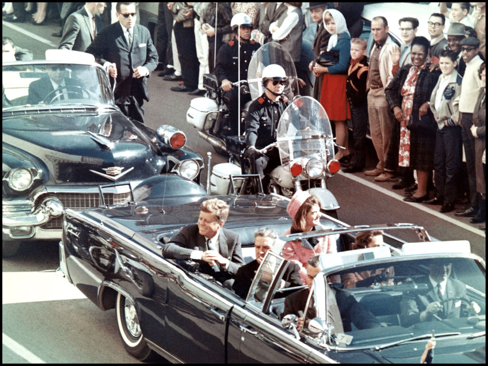 A car with president john f. Kennedy in the back driving past a motorcycle cop and other people