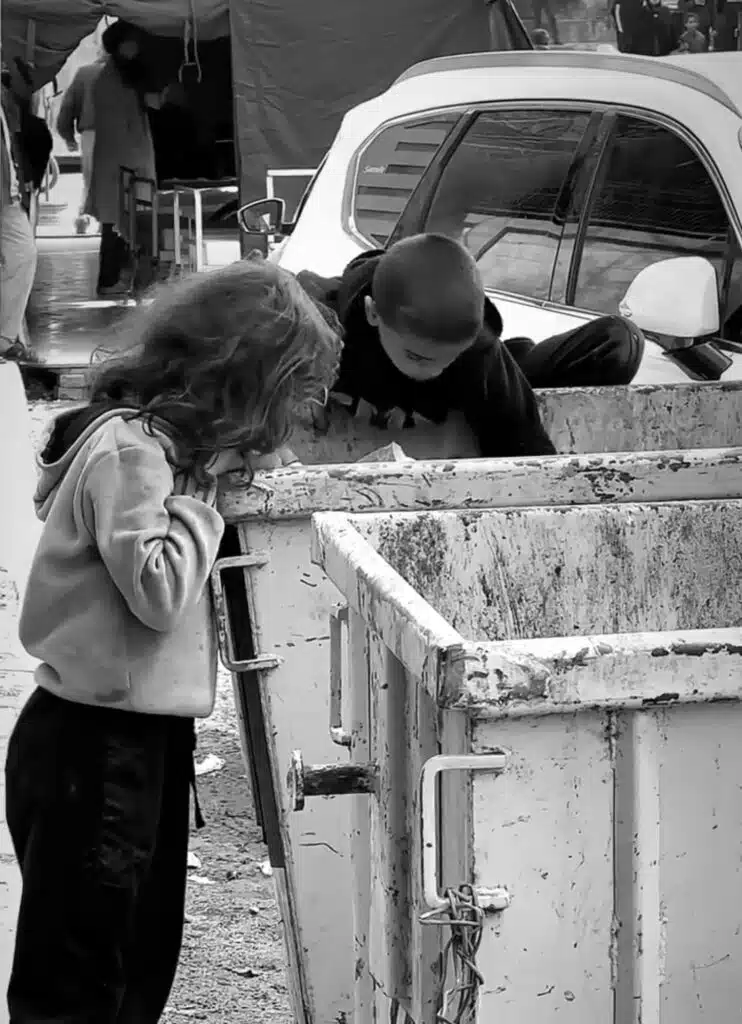 A woman standing next to a dumpster looking down at the ground.